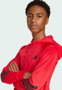 Sudadera atlética roja con collar de media cremallera, logotipo negro en el pecho y rayas negras en las mangas. Tela suave y ligera.