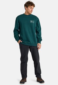 Turkusowy sweatshirt z okrągłym dekoltem i logo na przodzie, w połączeniu z czarnymi spodniami i brązowym obuwiem turystycznym. Gładka tekstura, luźny krój i długie rękawy.