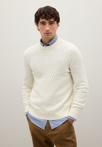 Cremefarbener Strickpullover mit strukturiertem Rautenmuster, rundem Ausschnitt und gerippten Bündchen. Kombiniert mit einem blauen Hemd mit Kragen darunter.