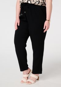 Zwarte elastische taillebroek met een losse pasvorm en zijzakken, gecombineerd met beige gevlochten hakken. Vlakke, gladde textuur.