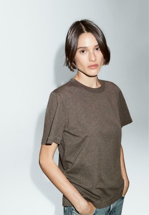 Brown cotton t-shirt with short, tapered sleeves and a round neck. Fabric appears soft with a smooth texture. Model stands against a light background.
