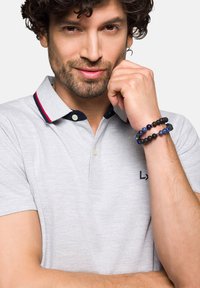 Jasnoszara koszulka polo z granatowymi i czerwonymi akcentami na kołnierzyku. Noszona z bransoletką na nadgarstek z wieloma koralikami w odcieniach ciemnych i niebieskich.