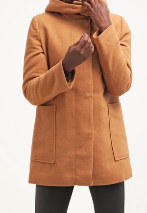 Personne portant un manteau en laine couleur camel avec de grandes poches avant, ajustant le col, sur un fond blanc uni.