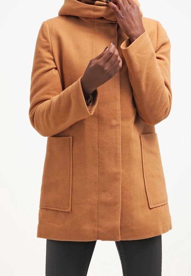 Personne portant un manteau en laine couleur camel avec de grandes poches avant, ajustant le col, sur un fond blanc uni.