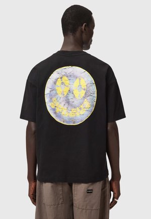 Mand vendt væk iført sort t-shirt med stort, revnet smiley-ansigt og gul tekst på ryggen, kombineret med brune bukser.