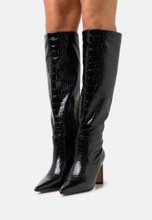 Bottes noires montantes en cuir verni avec une texture crocodile, bouts pointus et talon fin et haut. Détail zippé sur le côté.
