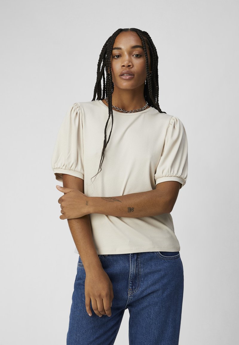 Lys beige ribbet t-shirt med puffede korte ærmer og rund halsudskæring, parret med blå denimjeans. Minimalistisk design uden synlige mønstre.