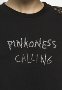 Sudadera negra con el texto "PINKONESS CALLING" contorneado en piedras de imitación claras. El escote es redondeado con detalles de encaje en los hombros.
