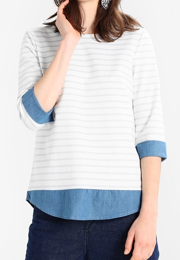 Femme portant un haut rayé blanc et gris clair avec des poignets et un ourlet en jean bleu, assorti à un jean bleu foncé, main près du visage.