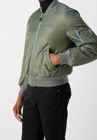 Veste bomber verte avec fermeture éclair, poignets côtelés et logo. Comprend des poches latérales et une poche zippée sur le bras. Tissu lisse et brillant.