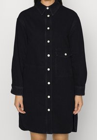 Mörk denimskjortklänning med krage, långa ärmar, vita knappar fram och bröstficka. Har en rak skärning med sidokli.