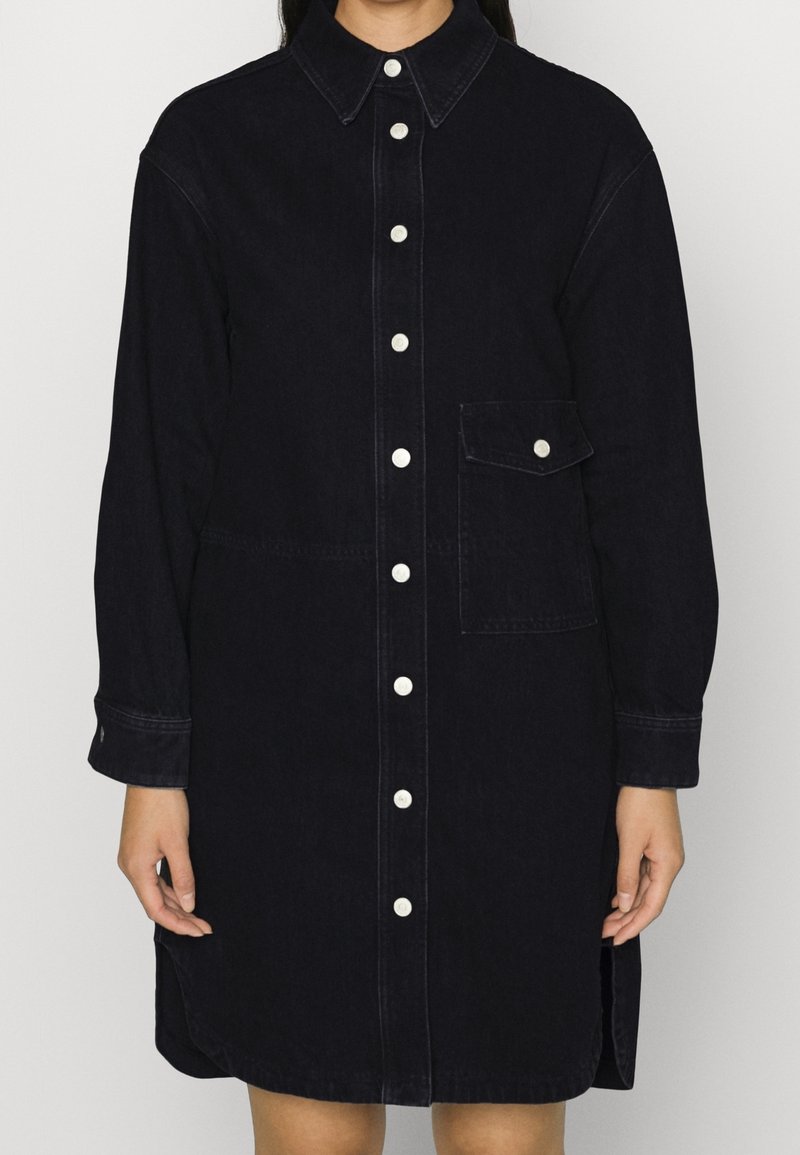 Mörk denimskjortklänning med krage, långa ärmar, vita knappar fram och bröstficka. Har en rak skärning med sidokli.
