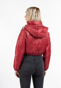 Giacca a vento rossa corta con cappuccio, caratterizzata da una texture lucida, polsini elasticizzati e un dettaglio con coulisse sul collo. Indossata sopra dei jeans scuri.