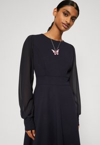 Merine dress, mille pikkade ja läbipaistvate varrukatega ning liibuva kehaosaga; kaunistatud roosa liblikakaelakeega hõbedasel kett. Sile kangas.