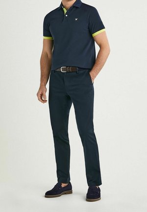 Polo-shirt navy con accenti verdi, abbinato a pantaloni scuri e scarpe scure. Caratterizzato da un colletto a due bottoni e un piccolo logo sul petto.