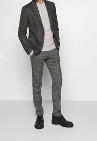 Grau karierte Hose kombiniert mit einem strukturierten schwarzen Blazer und einem hellbeigen Pullover. Schwarze Schnürschuhe runden das Outfit ab.