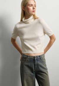 Suéter de punto de manga corta en color crema con cuello vuelto. Combinado con jeans de tiro alto en un azul desgastado que tienen cinco bolsillos y un ligero desgastado.