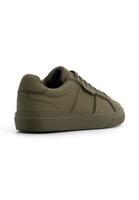 Sneaker verde oliva con superficie opaca liscia, dettagli testurizzati e lacci marroni. Presenta un profilo basso e una suola in gomma rigata.