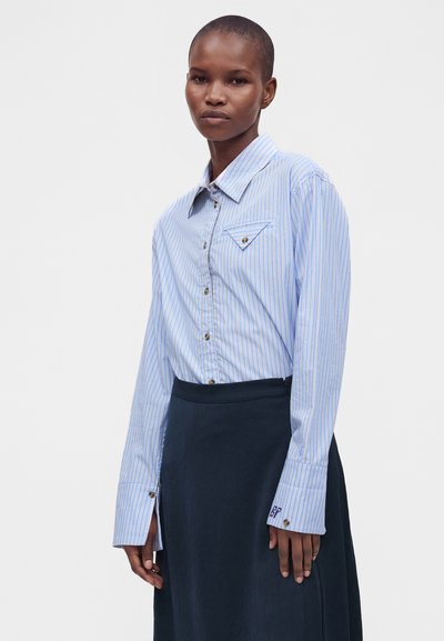 Femme portant une chemise à rayures bleu clair avec de larges poignets et une jupe foncée, debout devant un fond blanc uni.