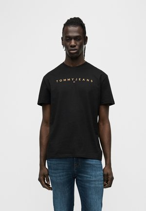 Muž stojaci pred jednofarebným pozadím, oblečený v čiernom tričku "TOMMY JEANS" a modrých džínsach, tvárou smerom vpred s neutrálnym výrazom.