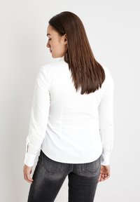 Weißes Hemd mit langen Ärmeln und abgerundetem Saum sowie seitlichen Schlitzen, kombiniert mit dunkelgrauen Jeans. Sichtbare glatte Stofftextur.