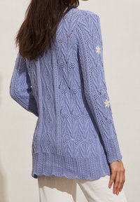 Světlé fialové pletené kardigan s výrazným vzorem copánků, zvlněným lemem a malým bílým květinovým detailem na rukávu.