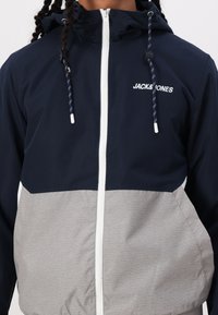 Marineblau und graue Windbreaker-Jacke mit Kapuze, ausgestattet mit einem weißen Reißverschluss und verstellbaren Kordeln sowie dem Logo "JACK & JONES" auf der Brust.