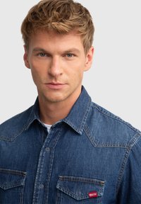 Denim krekls vidēji zilā krāsā ar klasisku apkakli, diviem krūšu kabatām un kontrastējošu šuvumu. Izcils sarkans logotipa uzlīme uz kreisās kabatas.