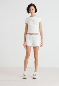 Witte, getailleerde t-shirt met een bloemenmotief en witte shorts met een structuurpatroon, gecombineerd met kleurrijke sneakers.