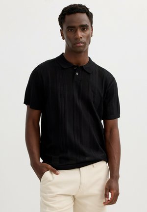 Hombre con una camisa polo negra de manga corta y texturizada y pantalones beige, de pie con una mano en el bolsillo frente a un fondo liso.