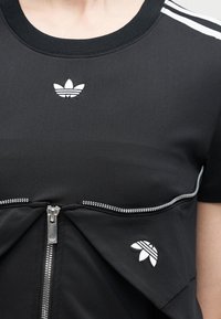 Μαύρο πουκάμισο Adidas με λευκά λογότυπα trefoil στο στήθος και στην πλευρά, με οριζόντιο ασημένιο φερμουάρ μπροστά.