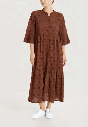 Donna che indossa un vestito midi marrone con bottoni, floreale con occhielli, maniche a campana 3/4 e sneakers bianche, in piedi davanti a uno sfondo semplice.