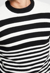 Pull rayé noir et blanc avec une texture en côtes, présentant des bandes horizontales de largeurs variées et un col rond.