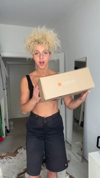 Caixa castanha da Zalando segurada por uma pessoa com cabelo loiro encaracolado, vestindo um top crop preto e calções pretos, de pé numa sala bem iluminada.