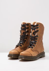Botas de cuero marrón con cordones y forro de pelo sintético, ojales negros y costuras amarillas en contraste en la suela de goma. Diseño grueso y texturizado.