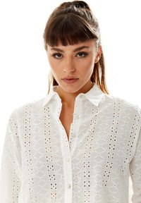 White button-up shirt featuring embroidered eyelet pattern, a collar, and a slightly textured surface. Visible button details on the front.