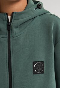 Zelená zipová mikina s texturovanou tkaninou, nastavitelnou kapucí s knoflíkem a černým logem na přední levé straně.