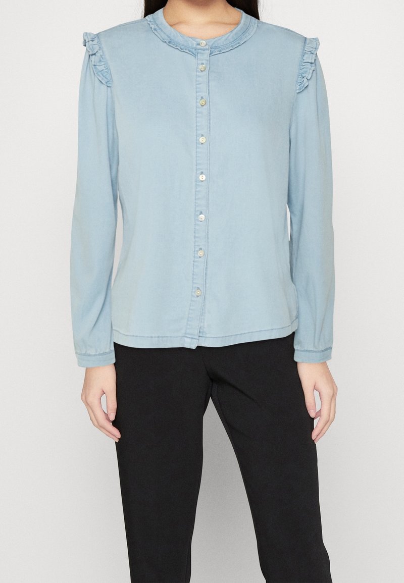 Femme portant un chemisier bleu clair boutonné avec des détails à volants sur les épaules et des manches longues, associé à un pantalon noir.