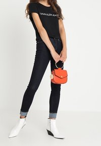 Svart t-shirt med tryck av "Calvin Klein Jeans", mörka jeans, vita ankelskor och en orange axelväska med spänne och cirkulär detalj.