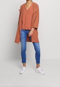 Camisa de color óxido con cuello en V y mangas holgadas, combinada con jeans ajustados azul y zapatillas blancas. Textura lisa, diseño casual.