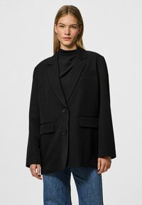 Blazer oversize nero con due tasche frontali e chiusura a singolo bottone. Texture liscia e design strutturato, abbinato a un top nero e denim.