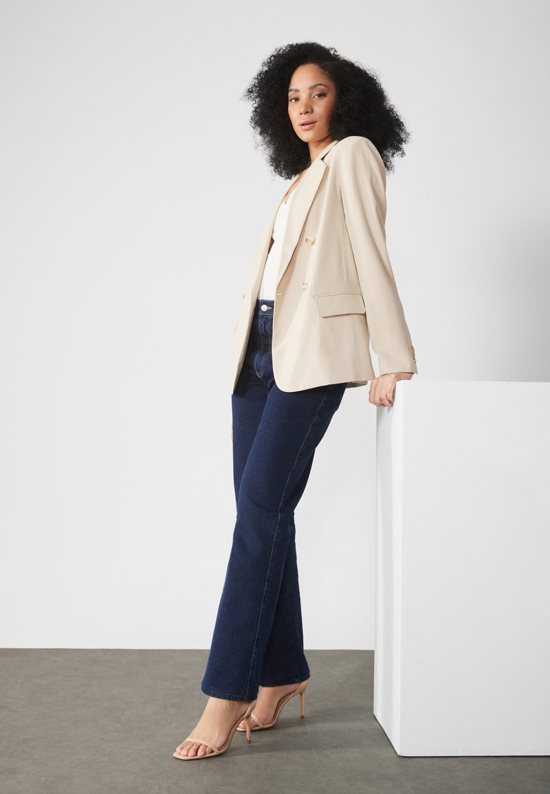 Blazer beige con bottoni dorati, top bianco e jeans a zampa d'elefante blu scuro. Modella in piedi accanto a un piedistallo bianco su una superficie grigia.