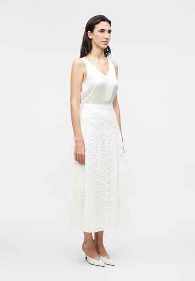 Γυναίκα που φοράει λευκό αμάνικο τοπ, λευκή midi φούστα με λουλουδάτη δαντέλα και λευκά μυτερά τακούνια μπροστά σε απλό φόντο.
