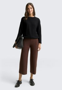 Schwarzer strukturierten Pullover, braune weite Hose mit vertikalen Nähten, schwarze Handtasche und schwarze Schnürschuhe. Neutraler Hintergrund.