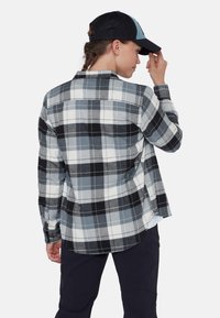 Camicia di flanella a quadri bianco e nero con accenti grigi, maniche lunghe e orlo arrotondato. Indossata con un berretto nero, che mostra un tessuto strutturato.