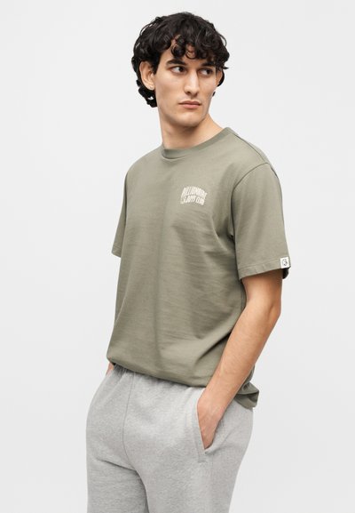 Λαδί κοντομάνικο μπλουζάκι με λογότυπο μπροστά. Απαλό βαμβακερό ύφασμα, άνετη φόρμα, στρογγυλή λαιμόκοψη, συνδυασμένο με ανοιχτό γκρι φόρμα.