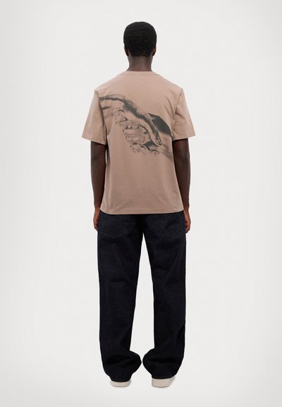 Braunes Baumwoll-T-Shirt mit einem schwarzen Grafikdesign von Händen auf der Rückseite, kombiniert mit dunkelblauen Jeans und getragen mit hellen Schuhen.