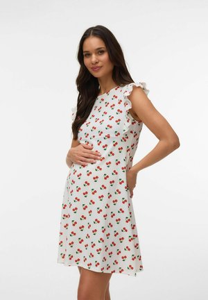 Tehotná žena s dlhými tmavými vlasmi, oblečená v bielych šatách bez rukávov s červeným motívom čerešní, drží si bruško a usmieva sa na jednoliatom pozadí.