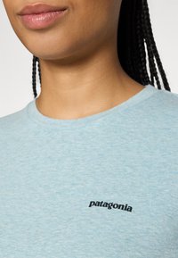 Tuvo tuvumā cilvēka, kas valkā gaiši zilu Patagonia T-kreklu, redzama logo uz krūšu un pīts matu daļa, kas krīt pāri plecam.
