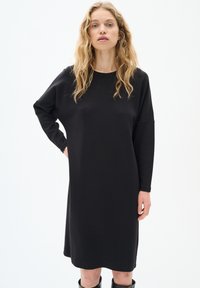 Juoda ilgam rankovėm suknelė, pagaminta iš minkštos medžiagos, pasižyminti laisvu siluetu ir apvaliu iškirpimu, su kelio ilgio dizainu ir minimalizmo stiliumi.